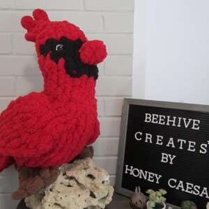 May include: A red crocheted cardinal bird plush with black facial markings around the eyes and beak, perched on a textured rock. Next to it is a black letter board with white text reading BEEHIVE CREATES BY HONEY CAESA.