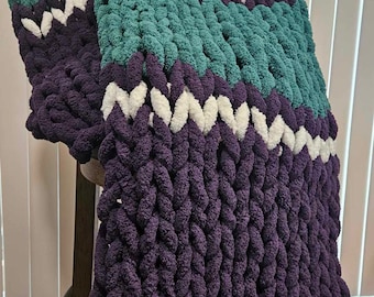 Könnte beinhalten: Eine dicke Strickdecke mit Streifen in Lila, Türkis und Weiß. Die Decke ist gefaltet und über einen Holzhocker gelegt.