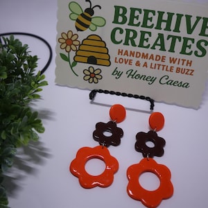 May include: Orange and brown flower-shaped earrings. The earrings have a three-tiered design with a circular top, a brown flower in the middle, and an orange flower at the bottom. The earrings are handmade by Honey Caeasa.