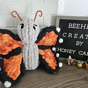 Puede incluir: Una mariposa de peluche con cuerpo gris, alas naranjas y negras y detalles blancos. La mariposa tiene antenas naranjas y negras. El letrero detrás de la mariposa dice "BEEHIVE CREATES BY HONEY CAESA".