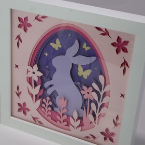 Könnte beinhalten: Gerahmtes, mehrschichtiges Papierkunstwerk mit Ostermotiv. Das Design zeigt eine Kaninchensilhouette in Eiform, umgeben von Blumen, Schmetterlingen und dekorativen Ausschnitten in Rosa, Lila und Weiß.