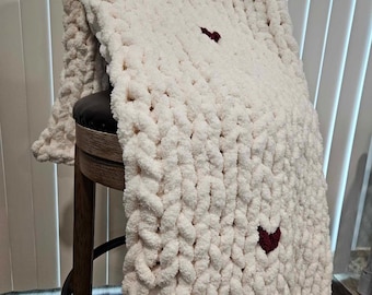 May include: A white chunky knit blanket with two burgundy hearts. The blanket is draped over a wooden stool.