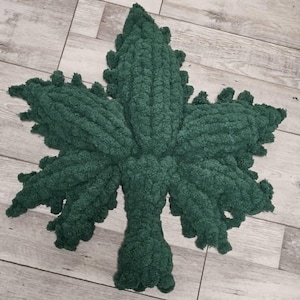 Op de afbeelding: Een groot, handgemaakt, donkergroen tapijt in de vorm van een blad. Het tapijt is gemaakt van een dik, getextureerd garen en het blad heeft gedetailleerde randen en nerven. Het tapijt ligt op een lichtgekleurde houten vloer.
