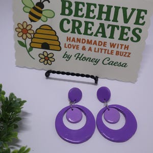 May include: A pair of purple, retro-style earrings with a circular design. The earrings are displayed in front of a sign that reads "BEEHIVE CREATES" with a bee and flower illustration. The sign also includes the text "HANDMADE WITH LOVE & A LITTLE BUZZ by Honey Caeasa."