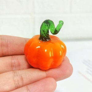 Pode incluir: Uma pequena figura de abóbora de vidro em laranja vibrante com uma superfície lisa e brilhante e um caule de vidro verde distintamente curvado. A abóbora tem uma forma arredondada e segmentada com cristas visíveis de vidro polido.