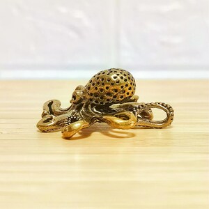 Puede incluir: Peque&ntilde;a y detallada figura de pulpo de lat&oacute;n. El pulpo tiene un cuerpo texturizado con m&uacute;ltiples agujeros y tent&aacute;culos rizados. La figura es de color marr&oacute;n dorado y est&aacute; sobre una superficie de madera clara.
