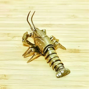 Pode incluir: Uma escultura metálica detalhada de um lagostim, com corpo segmentado, garras e antenas. O crustáceo está posicionado sobre uma superfície de madeira clara, mostrando seu design e textura intrincados. A cor da escultura é um dourado suave.