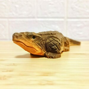 May include: A hand-carved wooden alligator figurine. The alligator is brown with detailed carvings, including scales and facial features. The figurine is resting on a light-colored wooden surface.