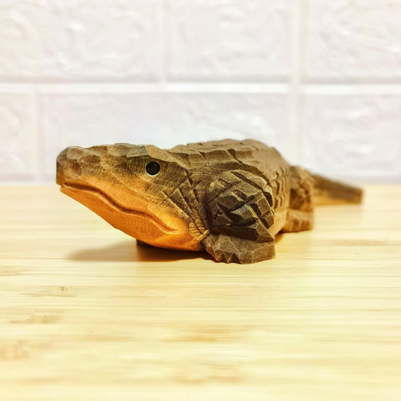May include: A hand-carved wooden alligator figurine. The alligator is brown with detailed carvings, including scales and facial features. The figurine is resting on a light-colored wooden surface.