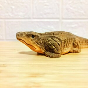 May include: A detailed wooden carving of an alligator, showcasing intricate textures and patterns. The sculpture is in a natural wood tone, with visible grain and carved details. The alligator is positioned on a light-colored wooden surface.