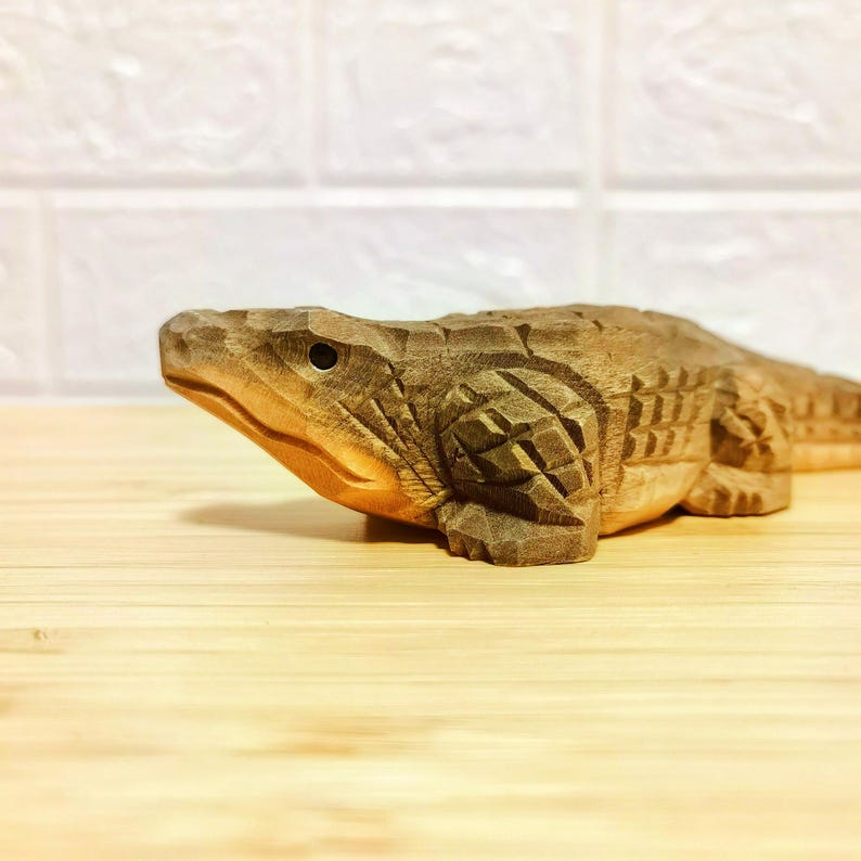 May include: A detailed wooden carving of an alligator, showcasing intricate textures and patterns. The sculpture is in a natural wood tone, with visible grain and carved details. The alligator is positioned on a light-colored wooden surface.
