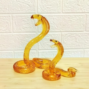 May include: Two amber glass cobra ornaments. The snakes are depicted with open mouths and raised heads, with detailed scale patterns. The figurines are on a light surface, set against a white brick wall.