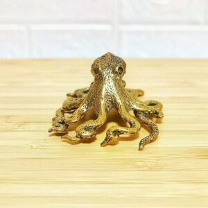 Puede incluir: Una detallada figura de pulpo de lat&oacute;n con piel texturizada y m&uacute;ltiples tent&aacute;culos. La escultura es de color dorado y se encuentra sobre una superficie de madera marr&oacute;n claro. El pulpo es un objeto decorativo.