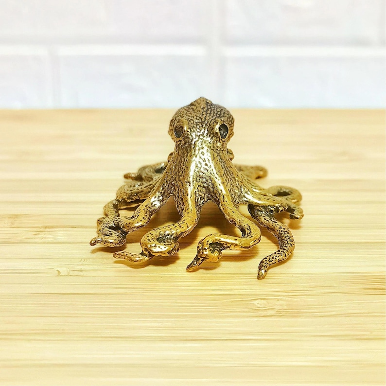 Puede incluir: Una detallada figura de pulpo de lat&oacute;n con piel texturizada y m&uacute;ltiples tent&aacute;culos. La escultura es de color dorado y se encuentra sobre una superficie de madera marr&oacute;n claro. El pulpo es un objeto decorativo.