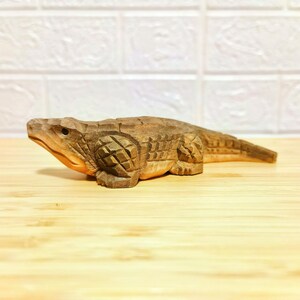 May include: A hand-carved wooden alligator figurine. The alligator is brown with detailed carvings, including scales and textured skin. The figurine is displayed on a light-colored wooden surface.
