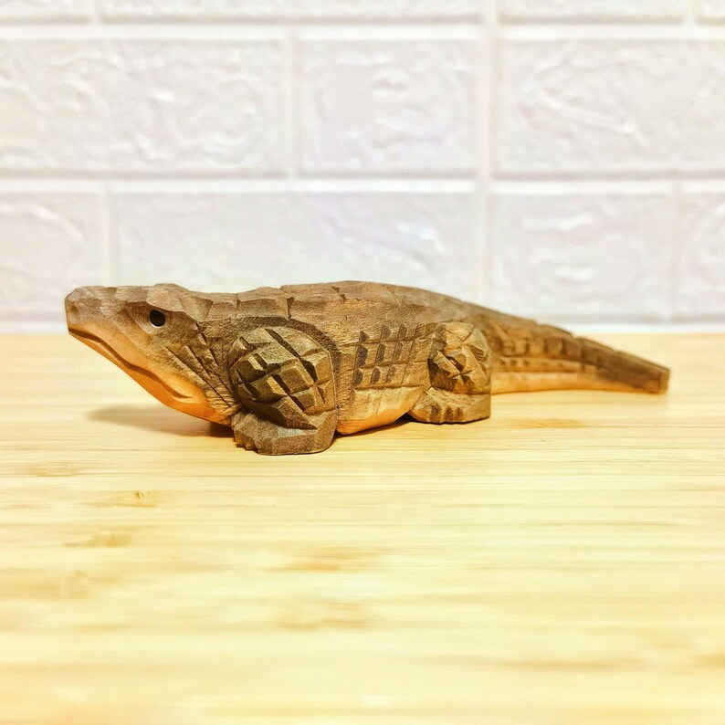 May include: A hand-carved wooden alligator figurine. The alligator is brown with detailed carvings, including scales and textured skin. The figurine is displayed on a light-colored wooden surface.