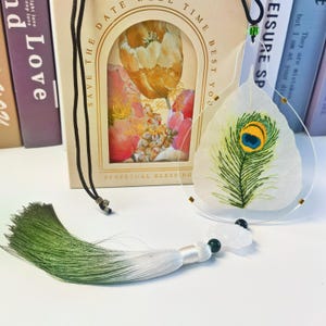 May include: A white leaf with a green peacock feather embroidered on it, framed in a clear plastic circle. The leaf is hanging from a black cord with a green tassel and a white jade pendant. The background is a white surface with a book in the background.