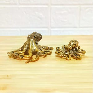 Puede incluir: Dos figuras de pulpo de lat&oacute;n. Una es m&aacute;s grande con un cuerpo s&oacute;lido, y la otra es m&aacute;s peque&ntilde;a con un cuerpo perforado. Ambas tienen tent&aacute;culos detallados y se exhiben sobre una superficie de madera clara.