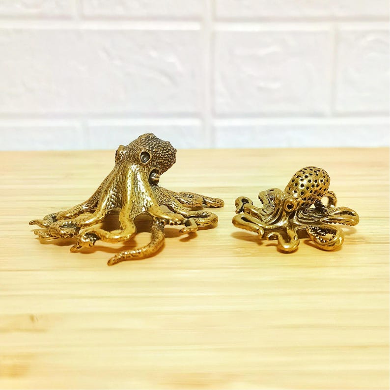 Puede incluir: Dos figuras de pulpo de lat&oacute;n. Una es m&aacute;s grande con un cuerpo s&oacute;lido, y la otra es m&aacute;s peque&ntilde;a con un cuerpo perforado. Ambas tienen tent&aacute;culos detallados y se exhiben sobre una superficie de madera clara.
