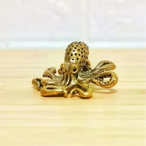 Puede incluir: Peque&ntilde;a y detallada figura de pulpo de lat&oacute;n. El pulpo tiene una cabeza texturizada con m&uacute;ltiples perforaciones y ocho tent&aacute;culos rizados. La figura es de color dorado y se encuentra sobre una superficie de madera clara.