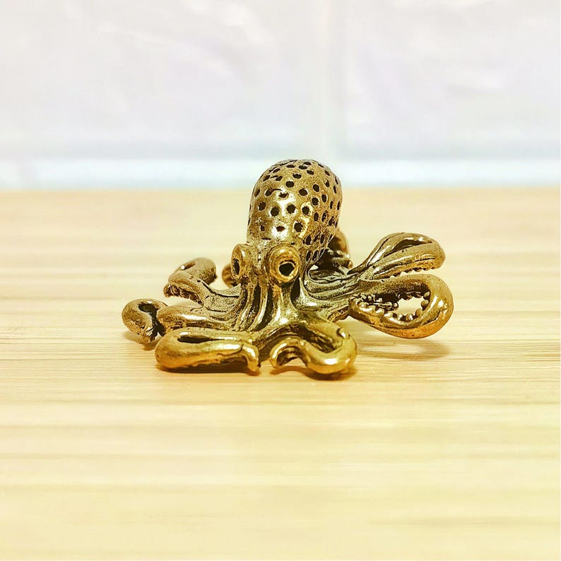 Puede incluir: Peque&ntilde;a y detallada figura de pulpo de lat&oacute;n. El pulpo tiene una cabeza texturizada con m&uacute;ltiples perforaciones y ocho tent&aacute;culos rizados. La figura es de color dorado y se encuentra sobre una superficie de madera clara.