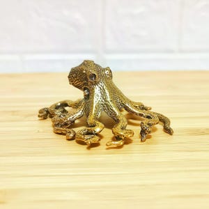 Puede incluir: Figura detallada de un pulpo de lat&oacute;n dorado, con piel texturizada y ocho tent&aacute;culos rizados. El pulpo es de color dorado y est&aacute; sobre una superficie de madera marr&oacute;n claro. Es un objeto decorativo.