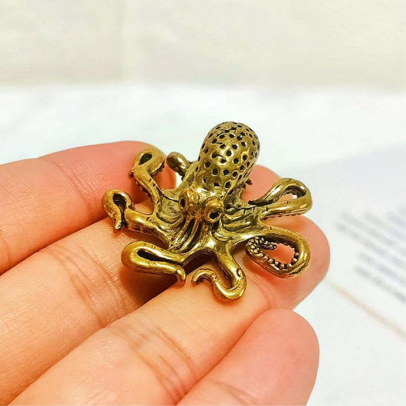 Puede incluir: Peque&ntilde;a y detallada figura de pulpo de lat&oacute;n. El pulpo tiene ocho tent&aacute;culos rizados y una cabeza texturizada con peque&ntilde;os agujeros. La figura es de color dorado c&aacute;lido y parece estar hecha de metal. El pulpo es un objeto decorativo.