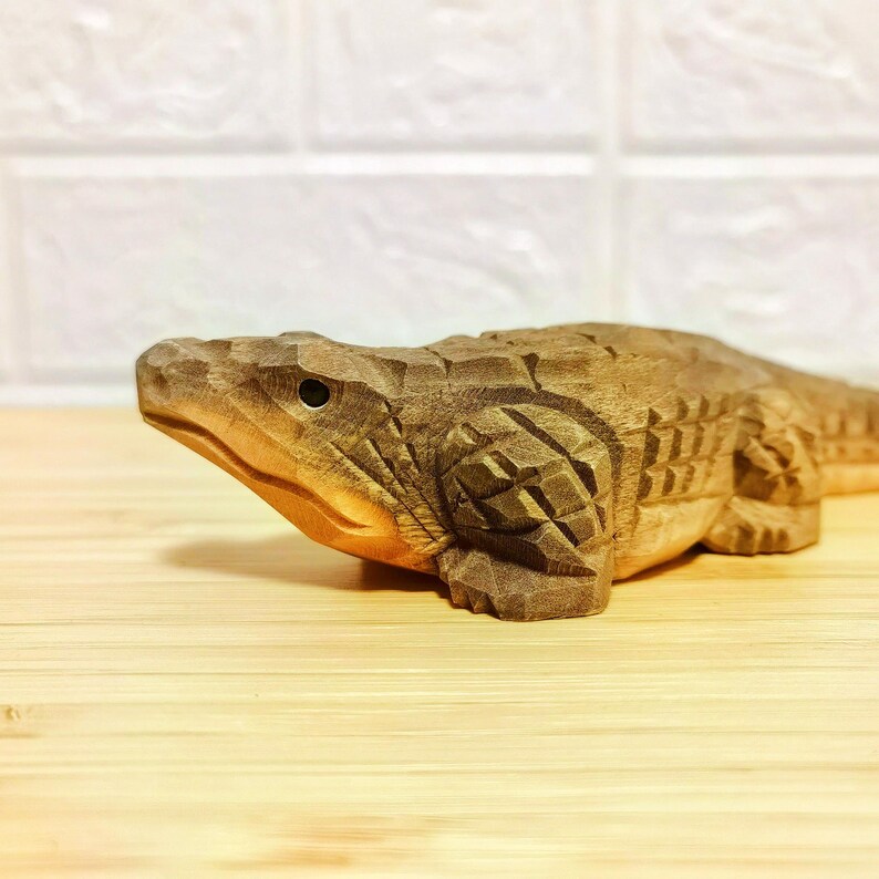 May include: A hand-carved wooden alligator figurine. The alligator is brown with detailed carvings, including scales and facial features. The figurine is lying on a light-colored wooden surface.
