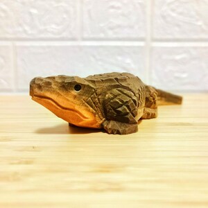 May include: A detailed wooden carving of an alligator, showcasing intricate textures and a natural brown hue. The alligator is positioned on a wooden surface, with a light-colored background. The carving highlights the alligator's scales and form.