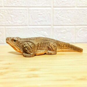 May include: A handcrafted wooden alligator figurine, carved with detailed scales and features. The alligator is a light brown color and rests on a wooden surface. The figurine is approximately 10 inches long.
