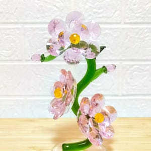 Peut inclure: Une sculpture florale en verre représentant des fleurs en cristal rose avec des centres jaunes et des tiges vertes. Les fleurs sont disposées en grappes, chaque pétale reflétant la lumière. La sculpture est posée sur un fond de briques blanches.