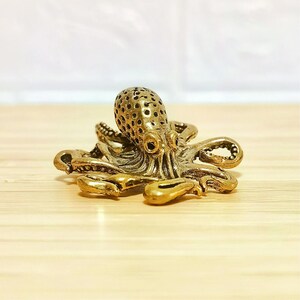 Puede incluir: Una detallada figura de pulpo dorada. El pulpo tiene una cabeza texturizada con peque&ntilde;os agujeros y m&uacute;ltiples tent&aacute;culos. La figura es de metal y se encuentra sobre una superficie de madera clara.