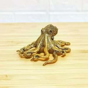 Puede incluir: Una detallada figura de pulpo dorada, con piel texturizada y ocho tent&aacute;culos extendidos. La escultura es de metal y se encuentra sobre una superficie de madera clara. El pulpo es un objeto decorativo.