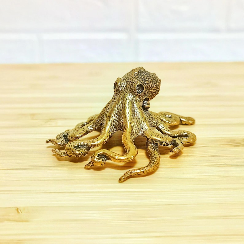 Puede incluir: Una detallada figura de pulpo dorada, con piel texturizada y ocho tent&aacute;culos extendidos. La escultura es de metal y se encuentra sobre una superficie de madera clara. El pulpo es un objeto decorativo.