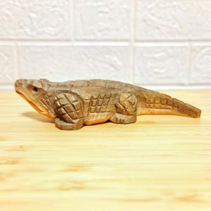 May include: A hand-carved wooden alligator figurine. The alligator is brown with detailed carvings on its body, legs, and head. The figurine is lying on a wooden surface. The alligator's mouth is slightly open, and its eyes are visible.