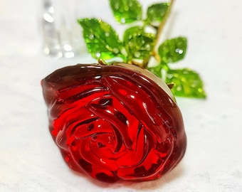 May include: A deep red glass rose flower with a smooth, rounded petal structure. The rose is attached to a thin, gold-toned stem featuring several vibrant green glass leaves with detailed veining and a glossy, reflective finish.