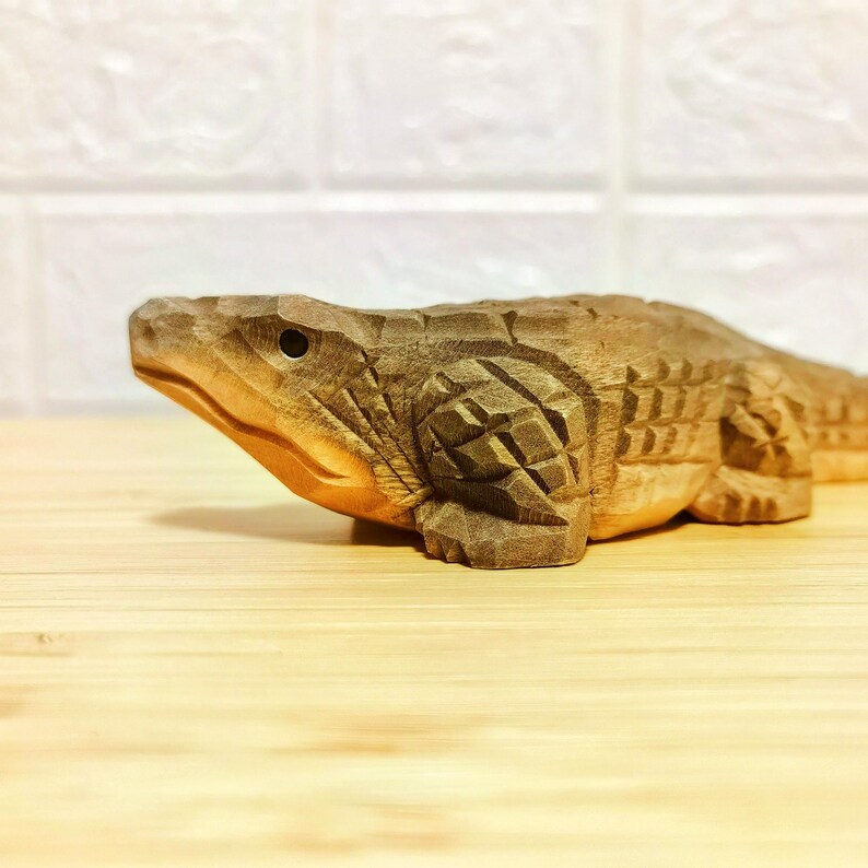 May include: A detailed wooden carving of an alligator, showcasing intricate textures and a natural wood finish. The sculpture is positioned on a light-colored wooden surface, with a white background.