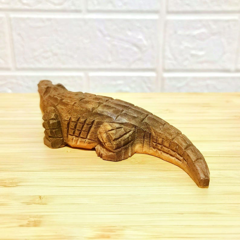 May include: A detailed wooden carving of an alligator, showcasing intricate textures and patterns. The alligator is brown, with visible scales and a textured body. The carving is placed on a light wooden surface.