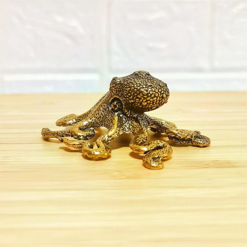 Puede incluir: Figura detallada de un pulpo de bronce con piel texturizada y tent&aacute;culos rizados. El pulpo es de color marr&oacute;n dorado y se encuentra sobre una superficie de madera clara. La figura es un objeto decorativo.