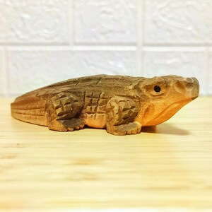 May include: A detailed wooden sculpture of an alligator, showcasing intricate carvings and a natural wood finish. The alligator is depicted in a lying down position, with visible scales and a detailed head. The sculpture is approximately 10 inches long.