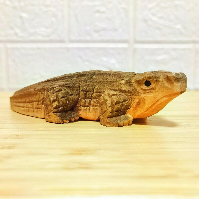 May include: A detailed wooden sculpture of an alligator, showcasing intricate carvings and a natural wood finish. The alligator is depicted in a lying down position, with visible scales and a detailed head. The sculpture is approximately 10 inches long.