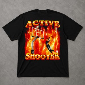 Può includere: T-shirt nero con grafica a tema basket. Il design presenta un giocatore di basket, un canestro e le parole "ACTIVE SHOOTER" in un font retrò con effetto fuoco. Lo sfondo presenta fiamme e fulmini, creando un effetto dinamico.