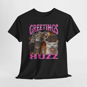 May include: Black t-shirt featuring a graphic of several cats with the text "GREETINGS HUZZ" in pink. The cats have various expressions, and the design has a retro, vintage aesthetic.