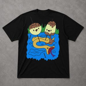 Puede incluir: Camiseta negra con un diseño de dibujos animados caprichoso. El gráfico representa dos personajes con forma de piruletas con glaseado de chocolate y vainilla, una serpiente amarilla y un fondo de agua azul. La serpiente tiene una lengua roja.
