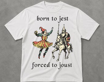 Puede incluir: Camiseta blanca con una ilustración medieval. El diseño muestra un bufón y un caballero a caballo, con las palabras "born to jest" y "forced to joust". El bufón lleva un traje colorido y el caballero está armado.