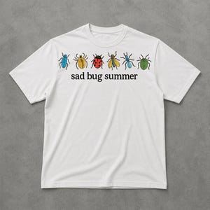 Może przedstawiać: Biała koszulka z rzędem kolorowych ilustracji owadów i napisem "sad bug summer". Owady są w odcieniach niebieskiego, żółtego, czerwonego i zielonego. Koszulka ma okrągły dekolt i krótkie rękawy.