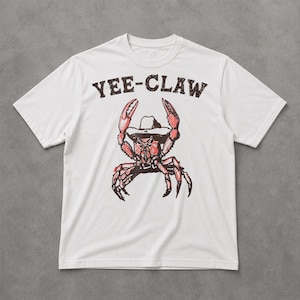 Op de afbeelding: Een wit T-shirt met korte mouwen en een afbeelding van een rode krab met een cowboyhoed op. Boven de krab staat de tekst YEE-CLAW in een zwart, verweerd lettertype. Het ontwerp is gecentreerd op de borst van het shirt.