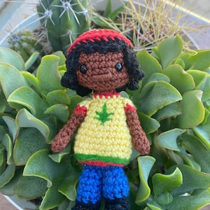Puede incluir: Muñeca de crochet con cabello negro, piel marrón y una camiseta amarilla con un diseño de hoja de marihuana verde. La muñeca lleva pantalones vaqueros azules y un gorro rojo.