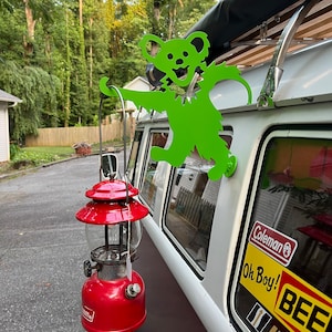 May include: A green metal cutout of a bear with a smiling face is attached to the side of a white van. A red Coleman lantern is hanging from the van's roof rack.