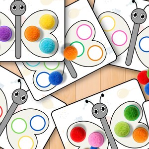 May include: A collection of butterfly-shaped educational cards with colorful pom-poms. Each card features a butterfly design with circles for pom-pom placement. The pom-poms are in various colors, including blue, purple, orange, and yellow.